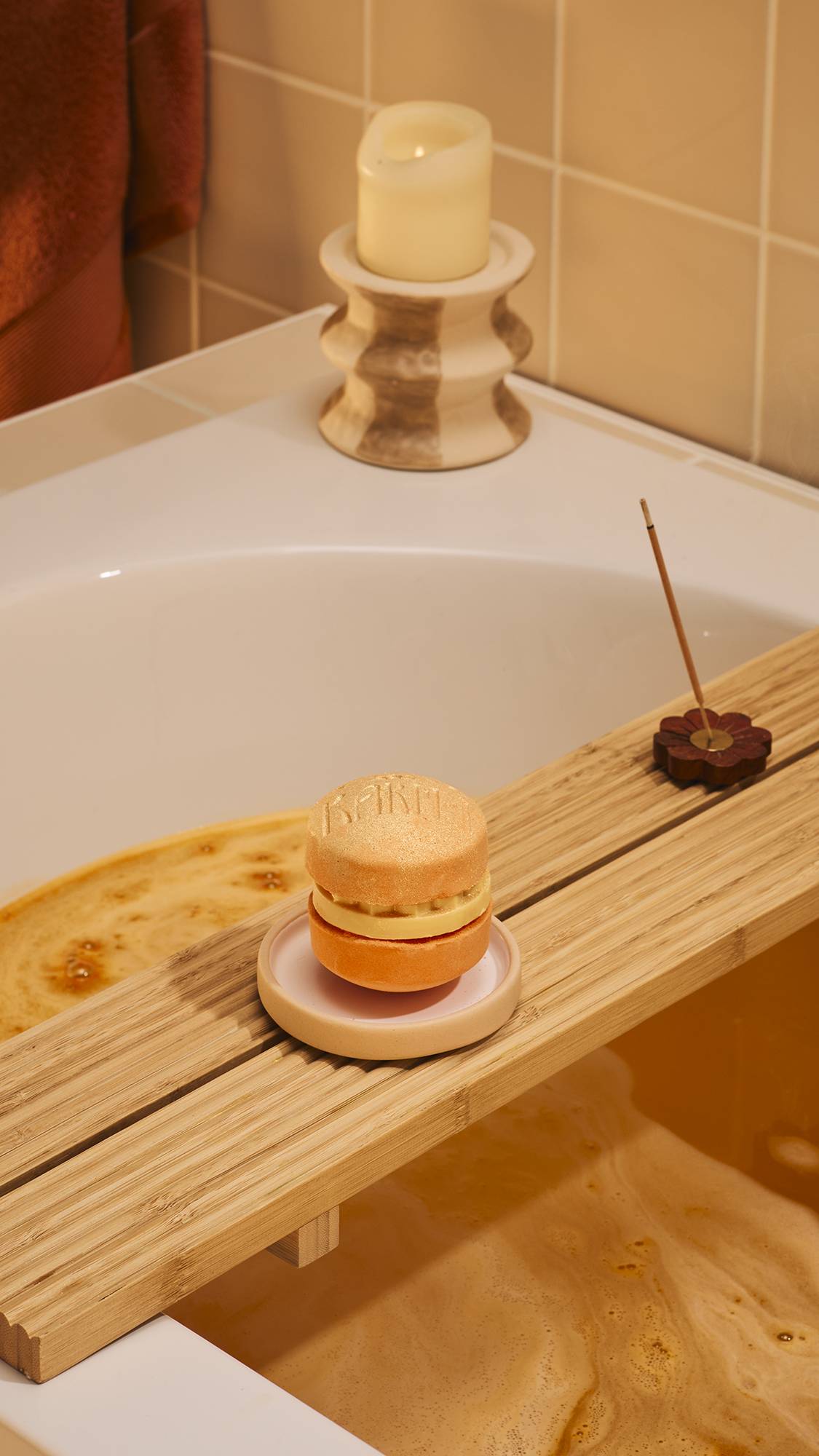 A shimmering orange 'Karma' macaron-style bath bomb sits on a small ceramic dish atop a wooden bath tray. In the background, a lit candle and a stick of incense provide a relaxing atmosphere against warm, beige bathroom tiles.