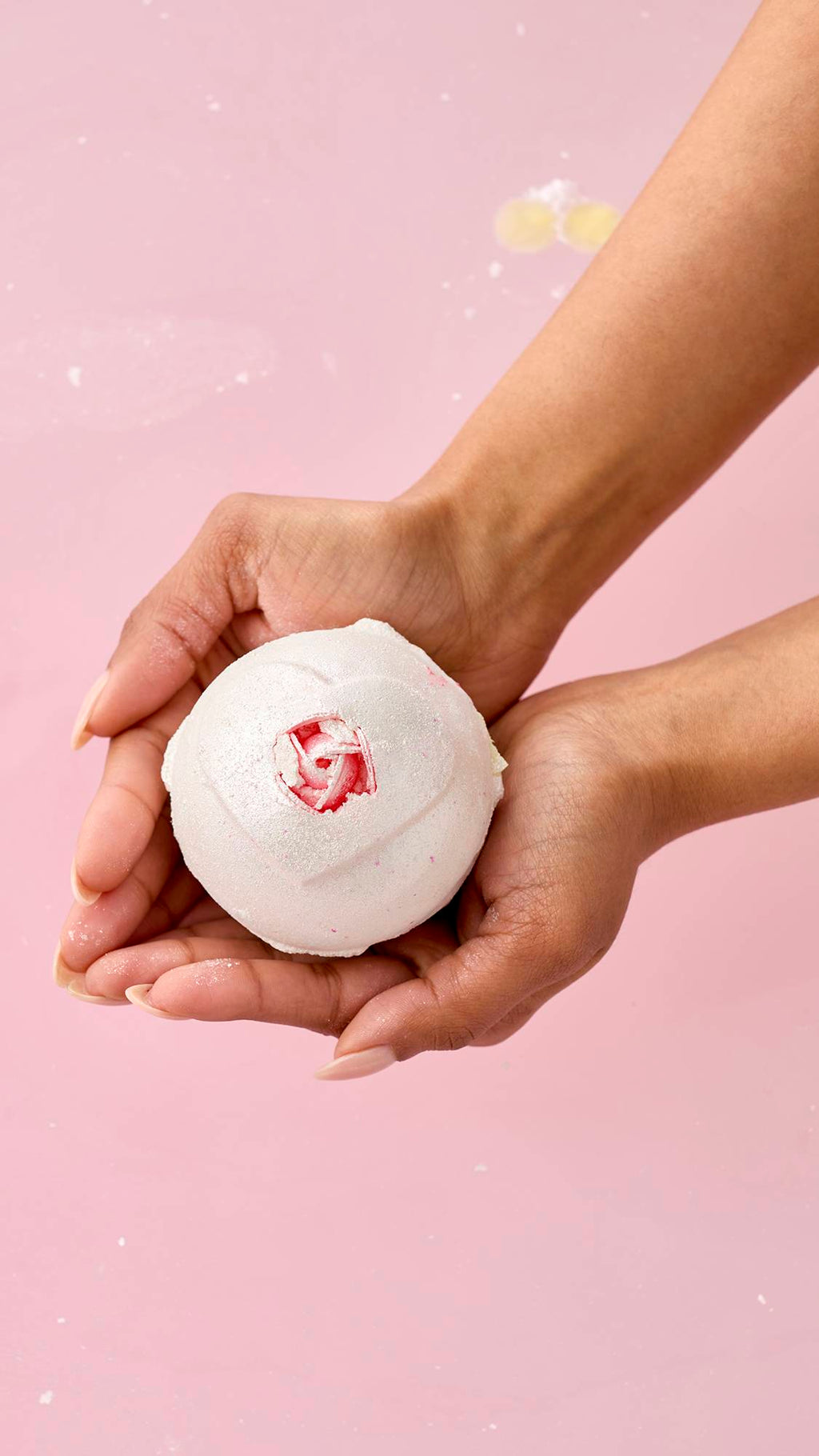 A pair of hands holds the pearlescent white Wedding bath bomb over pink bathwater. The sphere features an embossed heart shape with a small, realistic pink rosebud at its center.