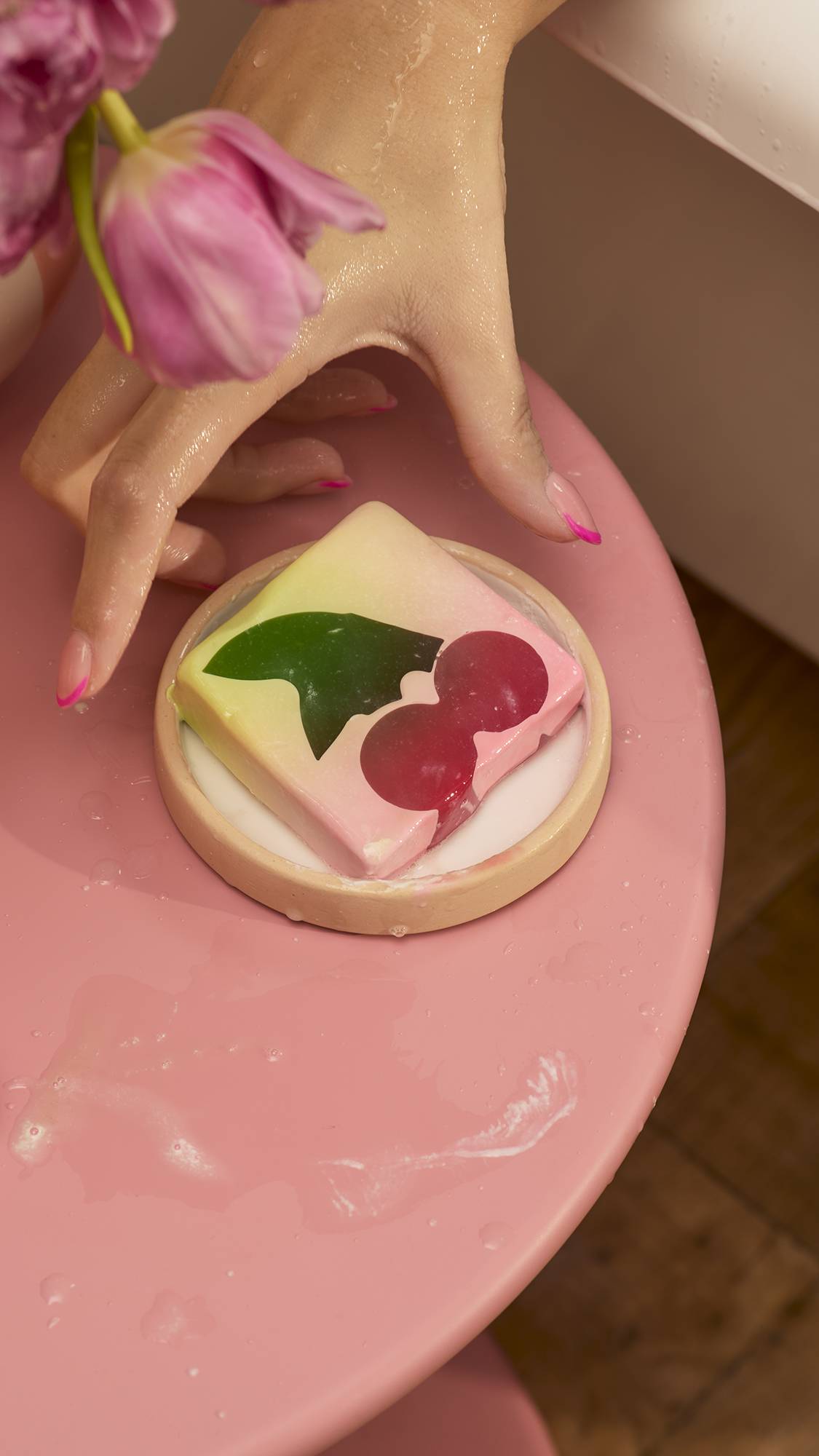 A square Cherry On Top soap bar with a dark cherry and green leaf design sits in a small dish on a pink table. A person with pink-tipped nails reaches for the soap near a white bathtub and a vase of pink flowers.