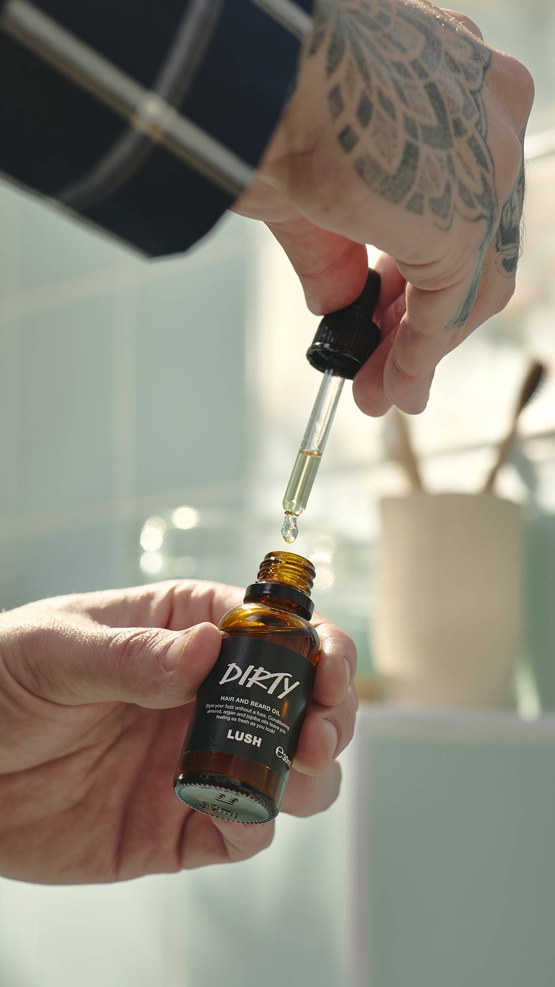 A model with a large geometric tattoo on their hand holds a small amber glass bottle of Lush "Dirty" hair and beard oil. They use a glass pipette to draw out a drop of the pale gold oil. Light green tiles are blurred in the background.