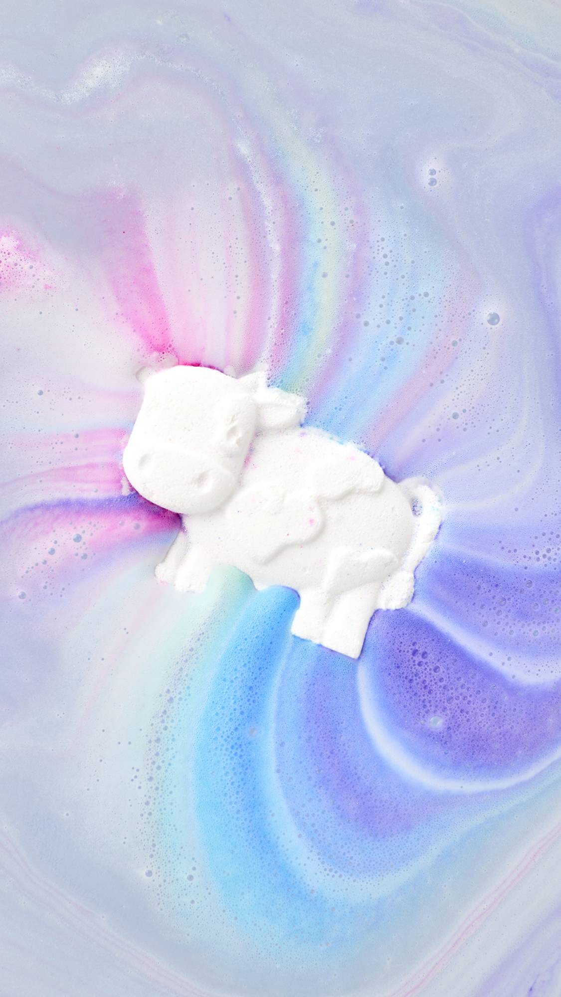 A top-down view of Toby's Magic Cow Bath Bomb as it dissolves in water, releasing a rainbow of colors. The white, cow-shaped bath bomb floats in the center, creating swirls of bright pink, blue, and purple fanning out across pastel purple bathwater.