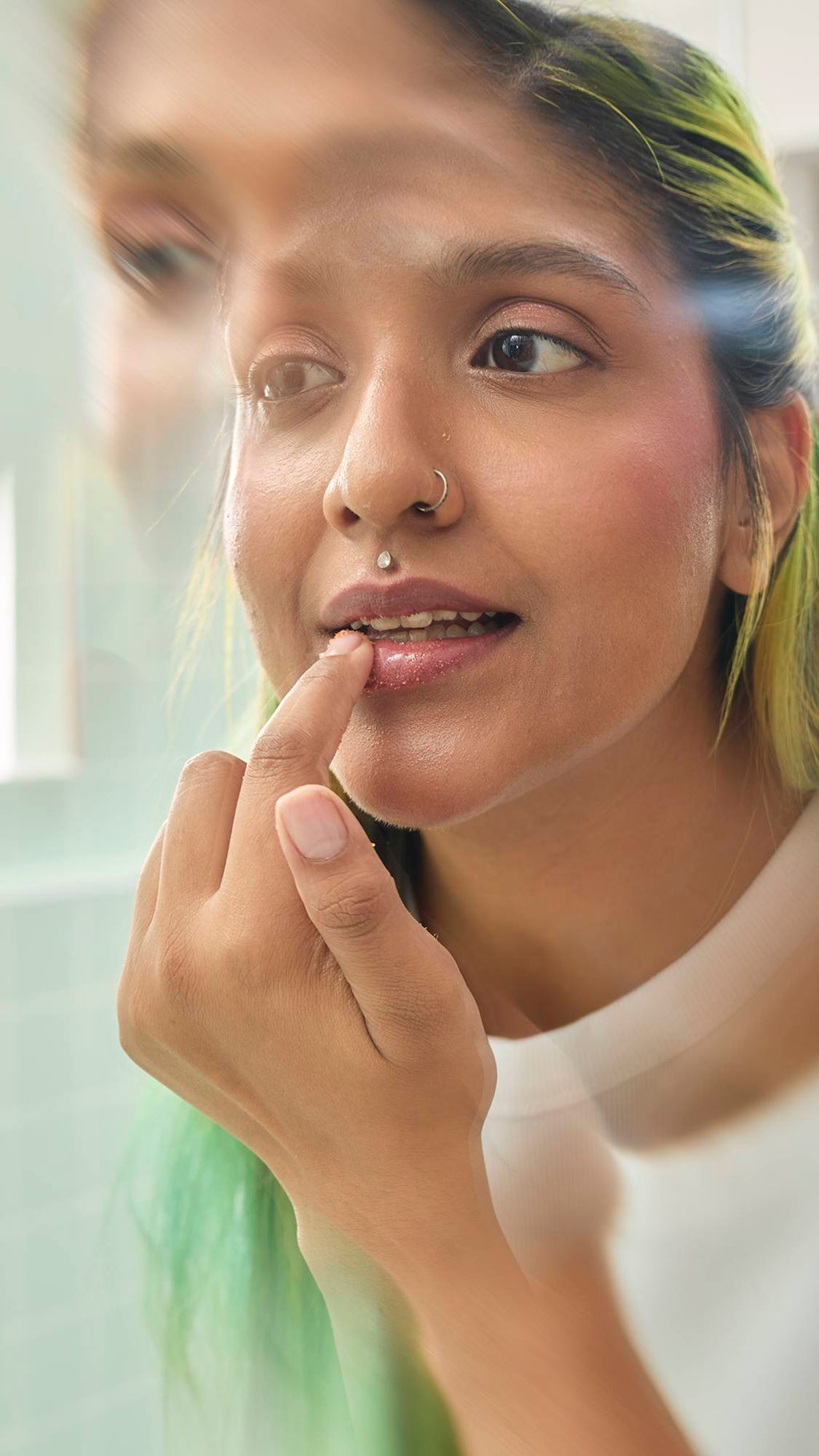 A close-up shot shows a model applying Lush "Buzz" Lip Scrub to their lips using their index finger. They have long, lime-green hair and facial piercings. The image is taken in a brightly lit bathroom, with a soft blur around the edges.