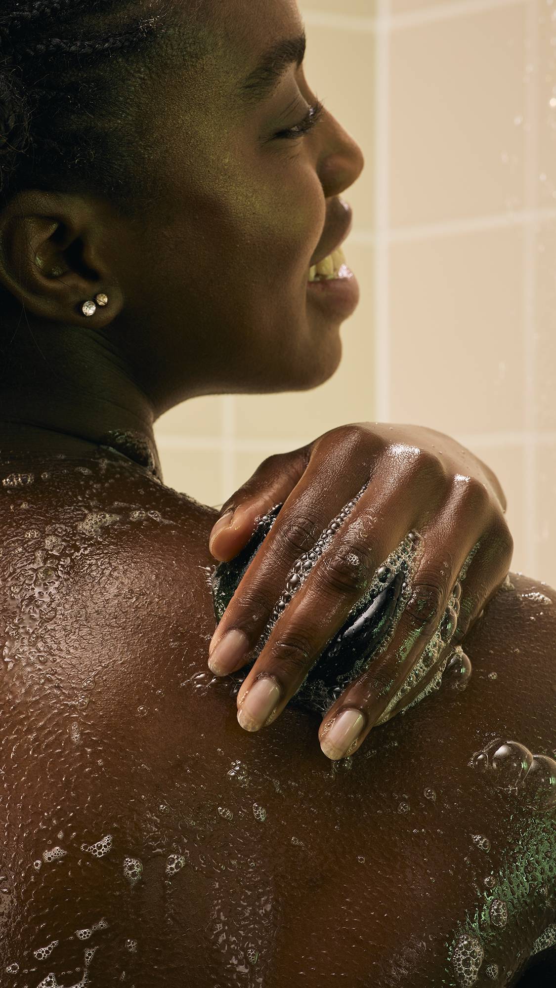A model with braided hair uses a dark green, translucent "The Good Hour" shower jelly to wash her shoulder. Water from the shower streams over her skin and the jelly, creating a glistening effect against the backdrop of neutral-toned bathroom tiles.