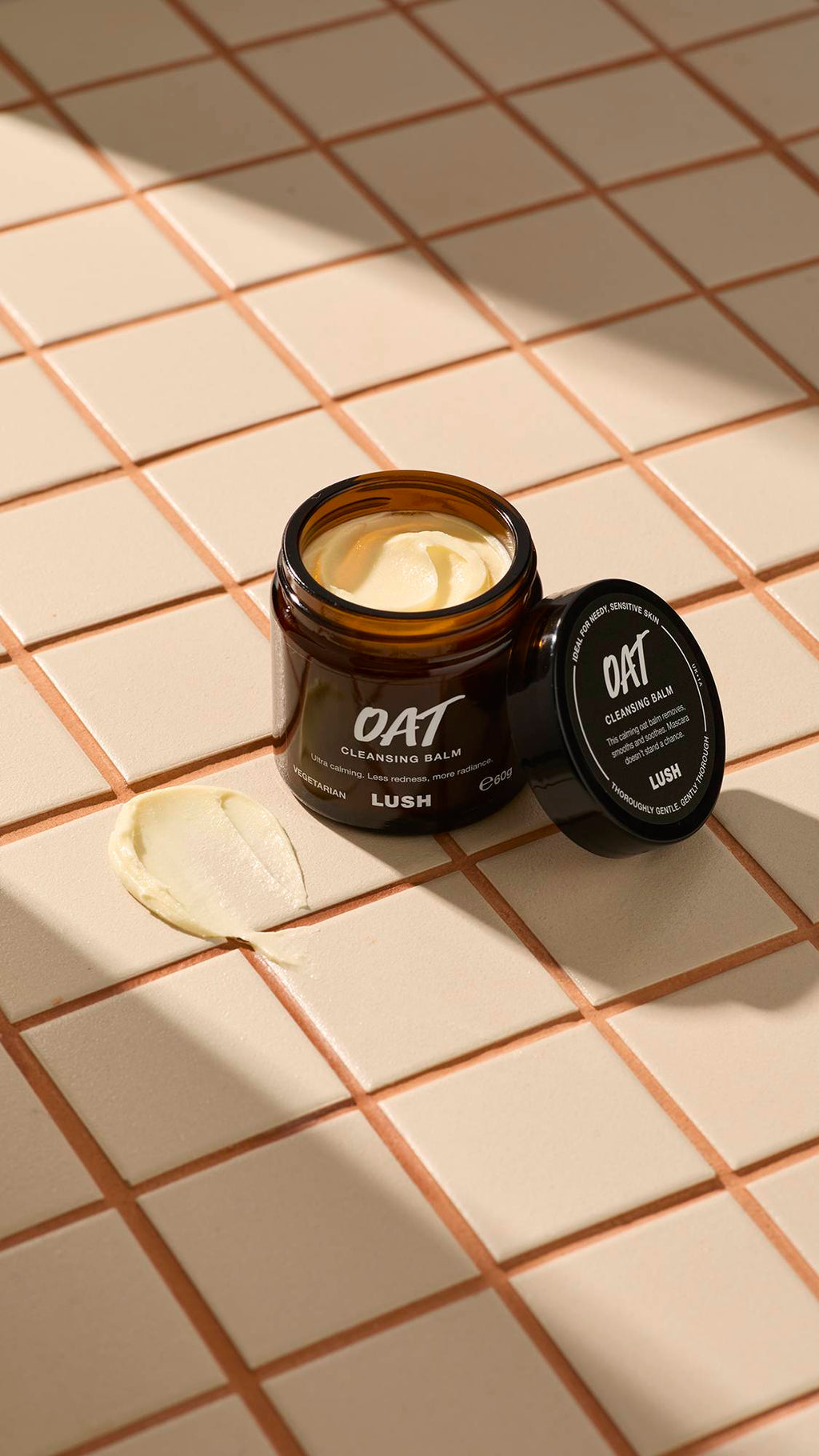 An open brown glass jar of Lush "Oat" facial cleanser sits on a beige tiled surface. A thick dab of the pale yellow, creamy product is smeared on the tiles next to the jar and its black lid.