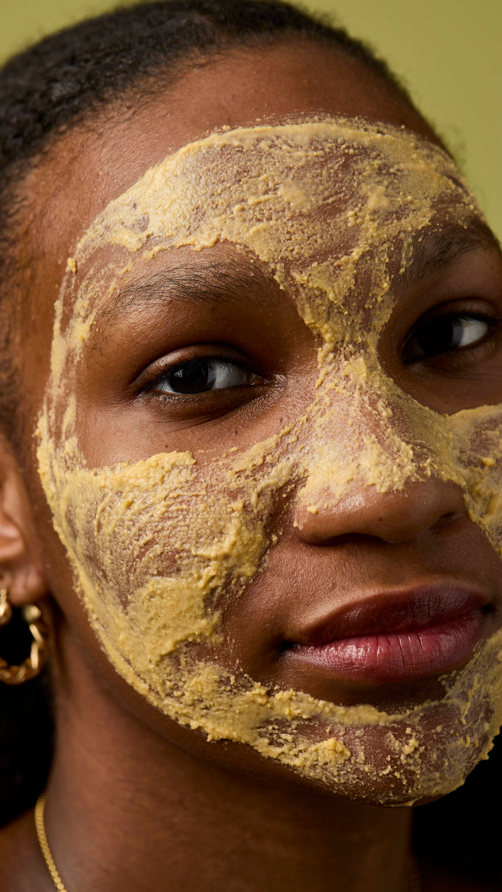 close-up of an individual with the thick, yellow, textured facial cleanser (Let the Good Times Roll) applied fully over their face, covering the forehead, cheeks, and nose. The background is a solid, muted olive-green colour