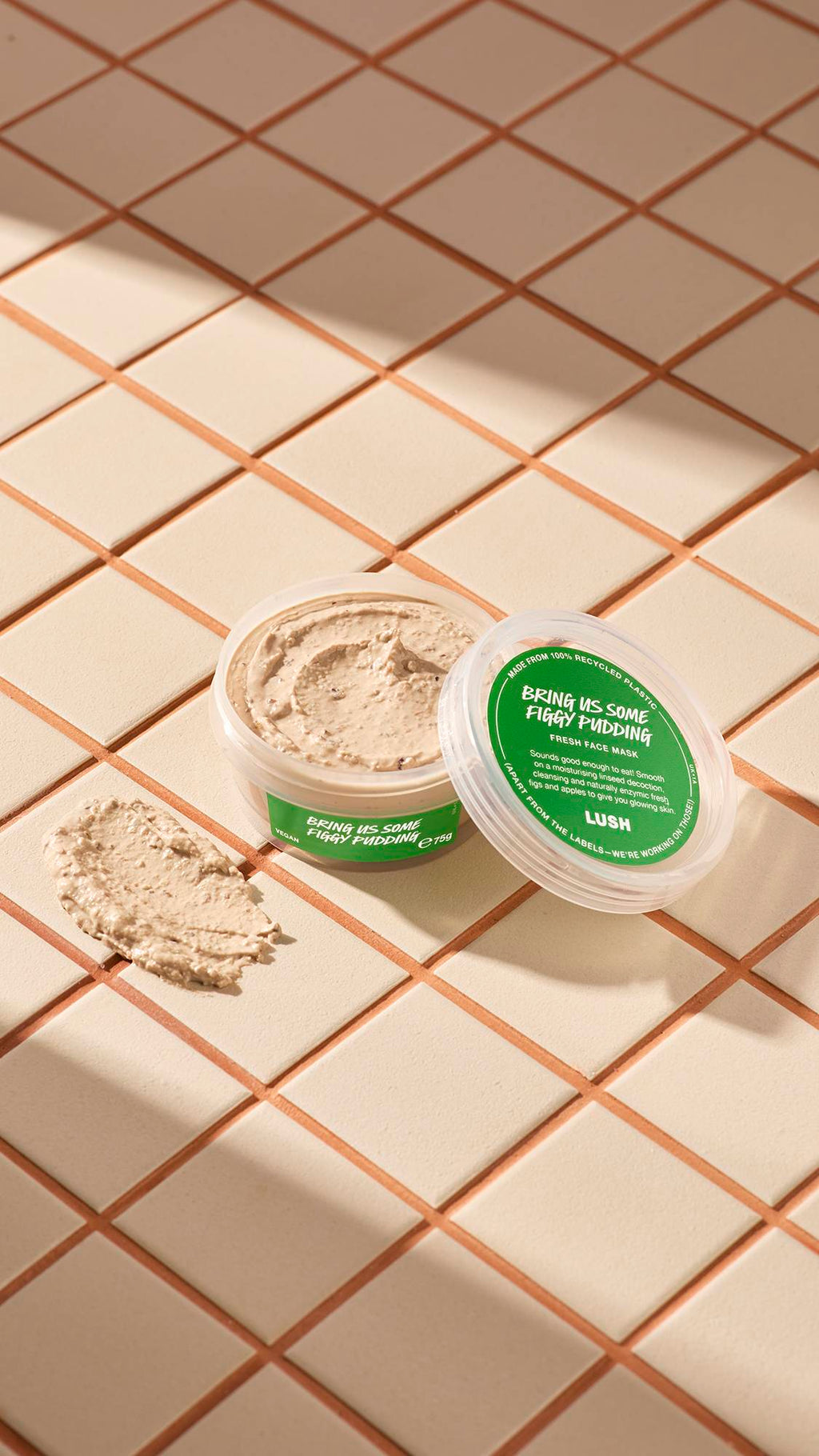 The image shows a clear, recycled plastic, open pot of Bring Us Some Figgy Pudding face mask with green labels, the lid leaning on the side and a smeared swatch of the textured product on the cream-tiled floor beside the pot.