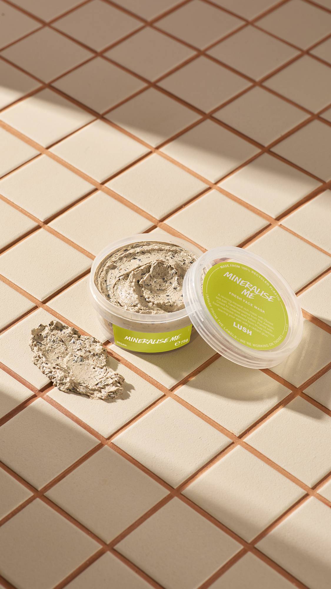 The image shows a clear, recycled plastic, open pot of Mineralise Me face mask with lime-green labels, the lid leaning on the side and a smeared swatch of the textured product on the cream-tiled floor beside the pot.