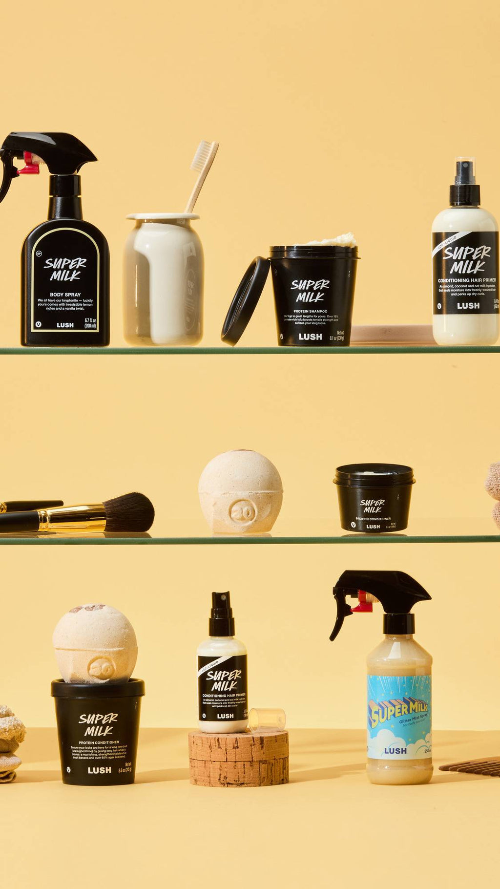 The LUSH Super Milk collection is arranged on glass shelves against a yellow background. It features the Body Spray, Hair Primer, Protein Shampoo, Conditioner, and Glitter Mist Spray in black and white packaging.