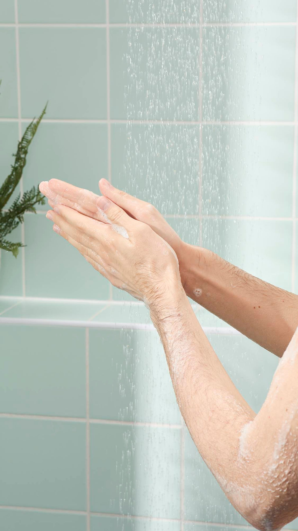 A close-up of a person’s arms and hands in a shower, lathering a light, soapy foam. The background features mint green wall tiles and the soft, vertical spray of water from a showerhead.