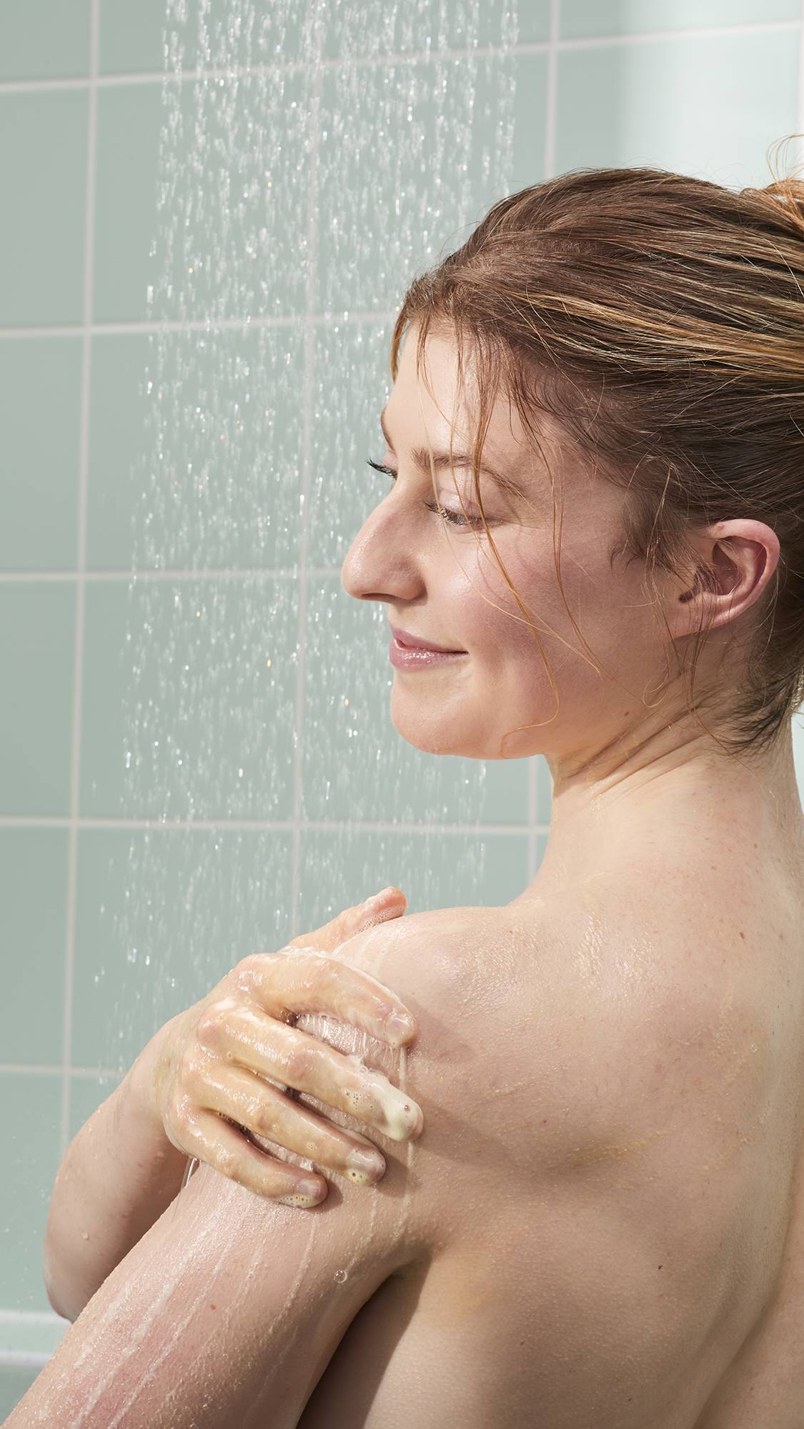 A woman with reddish-brown hair smiles in the shower while lathering a shimmering gold gel onto her shoulder. The background features light mint green tiles and the soft, vertical spray of falling water.