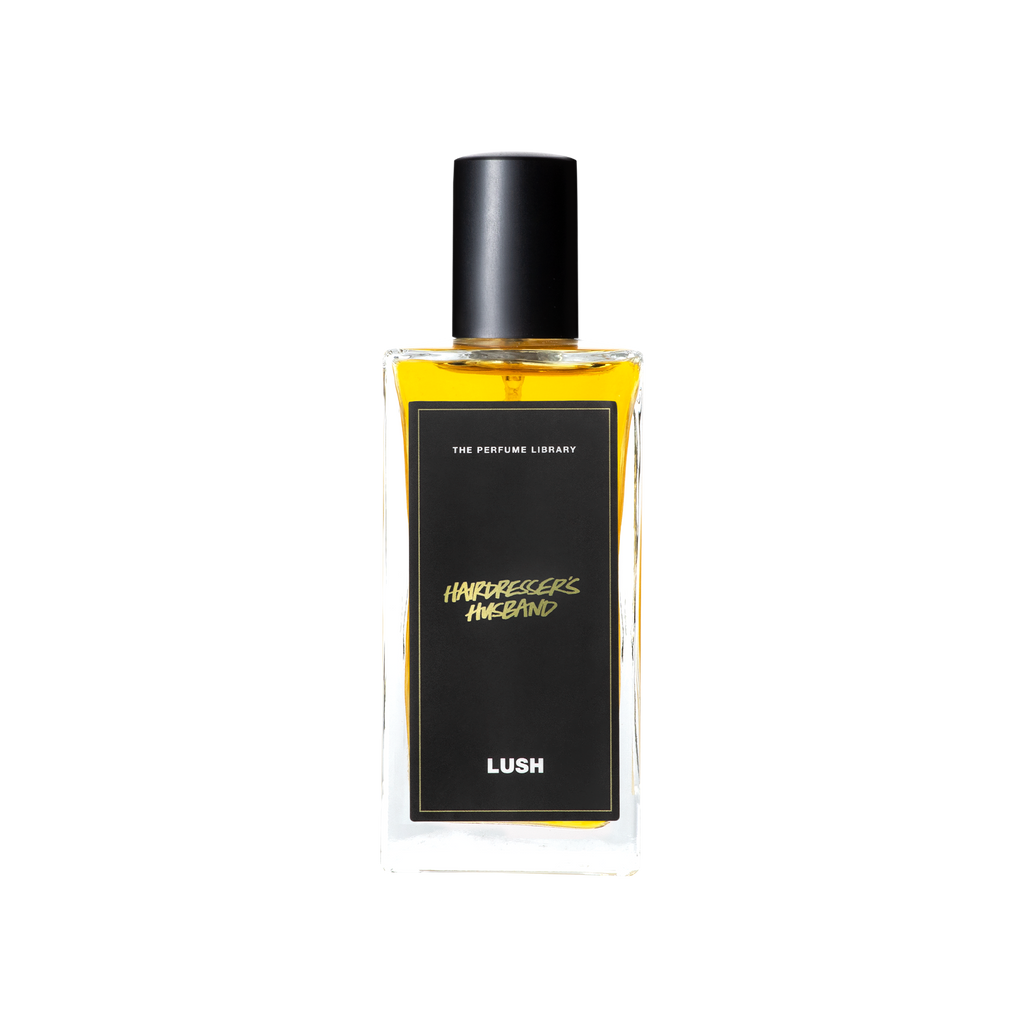 A glass perfume bottle filled with a light amber liquid. A black label reads 'The Perfume Library' and 'Hairdresser's Husband'.