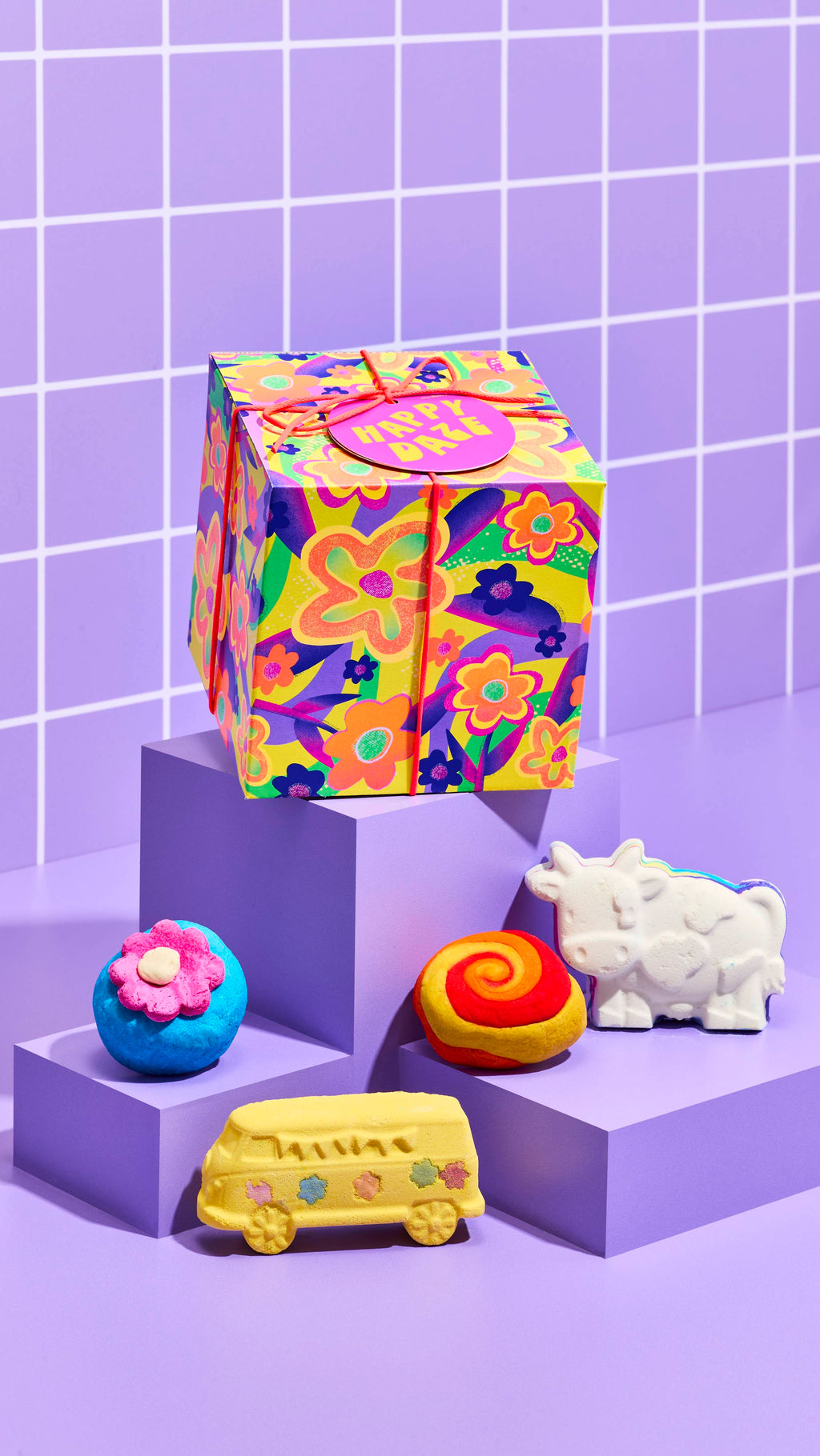 The image shows the colourful neon flower-designed Happy Daze gift box, sitting on a cubed platform with the four included bath products sitting on lower platforms on a pastel-purple tiled background.