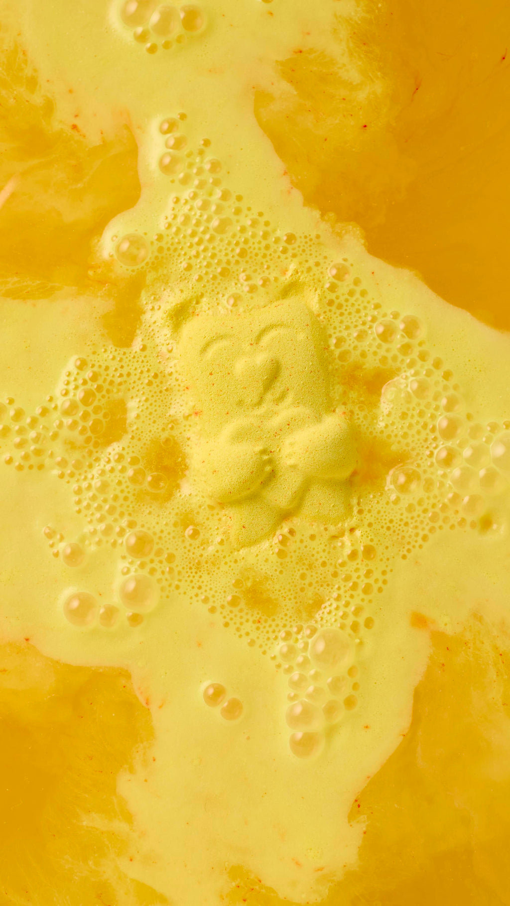 A close-up, top-down view of a yellow "Mama Bear" bath bomb dissolving in water. The bear fizzes in the centre, creating a bright yellow, foamy lather and bubbles that spread across the surface of the golden-yellow bathwater.