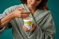 The image shows the model wrapped in a thick, sage-green dressing gown on a teal background. They are holding the white, LUSH version-themed packed pot of the Pineapple facial wash and scooping out the product with their other hand.