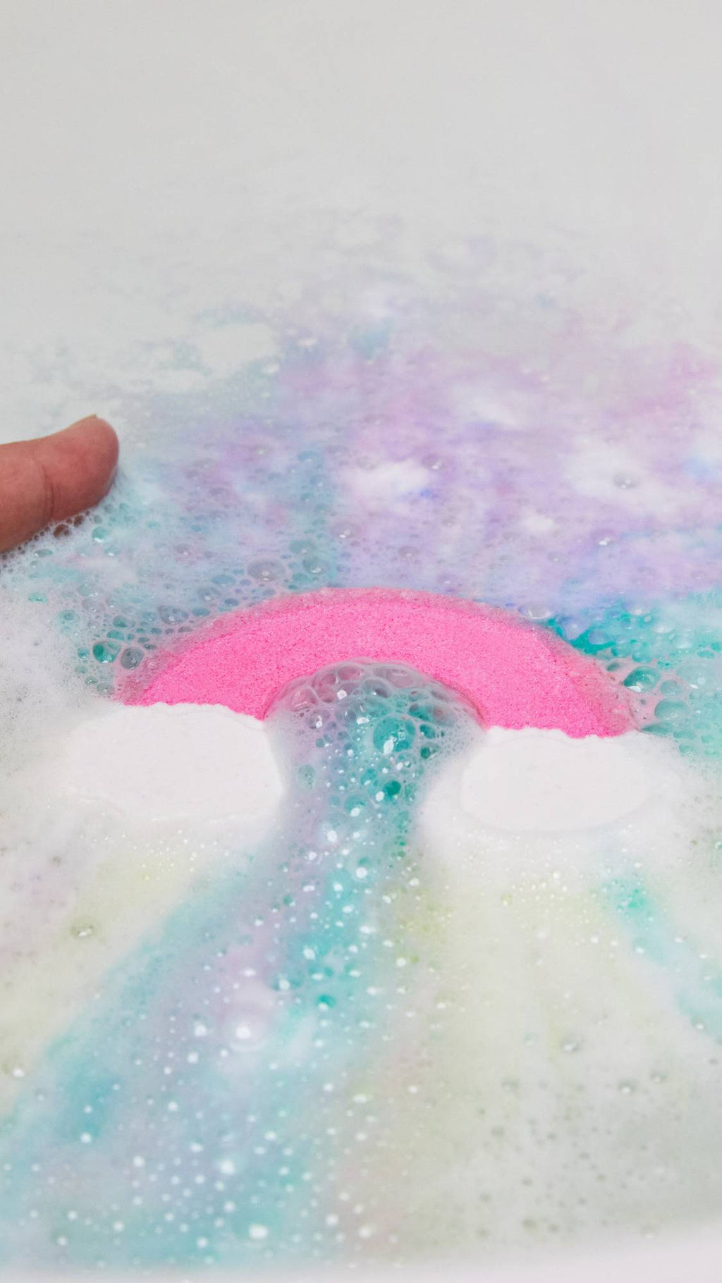 A close-up of the Somewhere bath bomb being placed into the bath water as it immediately ripples out pastel blue, pink and purple.
