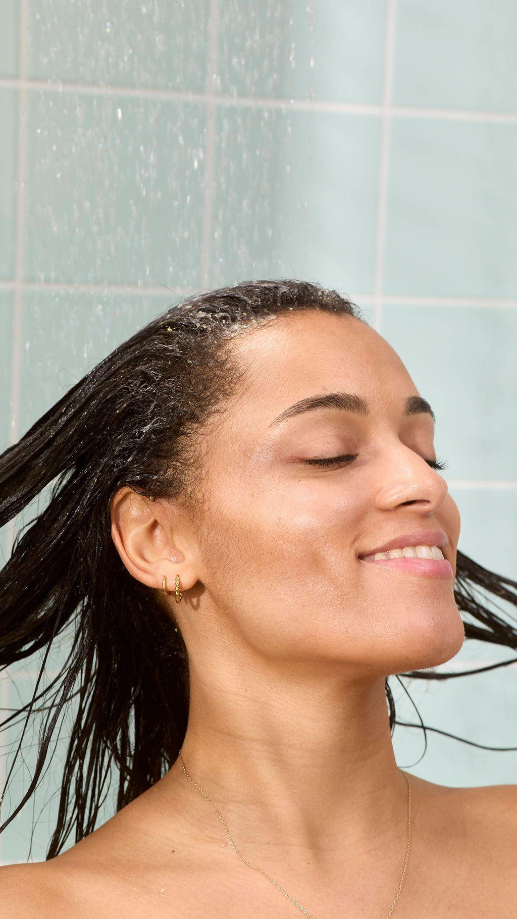 A model is smiling with their eyes closed while rinsing their dark hair under a shower. They are rinsing out the "St Clements" hair treatment product, set against a background of light green bathroom tiles with water droplets visible in the air.
