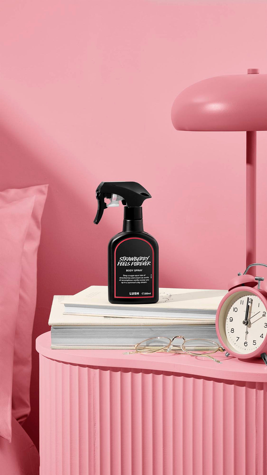 The image shows a recycled black plastic LUSH spray bottle of "Strawberry Feels Forever" sitting on a pile of books on a dusky pink bedside table. A matching coloured lamp, alarm clock, and glasses sit nearby, next to a pink bed and wall.