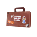 Suitcase. A brown cardboard gift box shaped like a suitcase with a carry handle. The front features 'PADDINGTON x LUSH' branding, an illustration of Paddington Bear in a towel, and a 'WANTED ON VOYAGE' sticker.